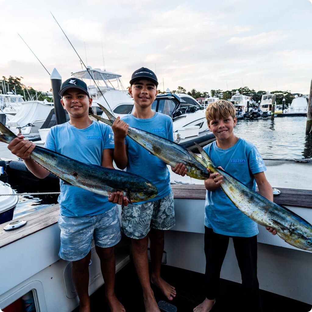 Three boys in blue shirts proudly holding large freshly caught fish on a boat docked at a marina.